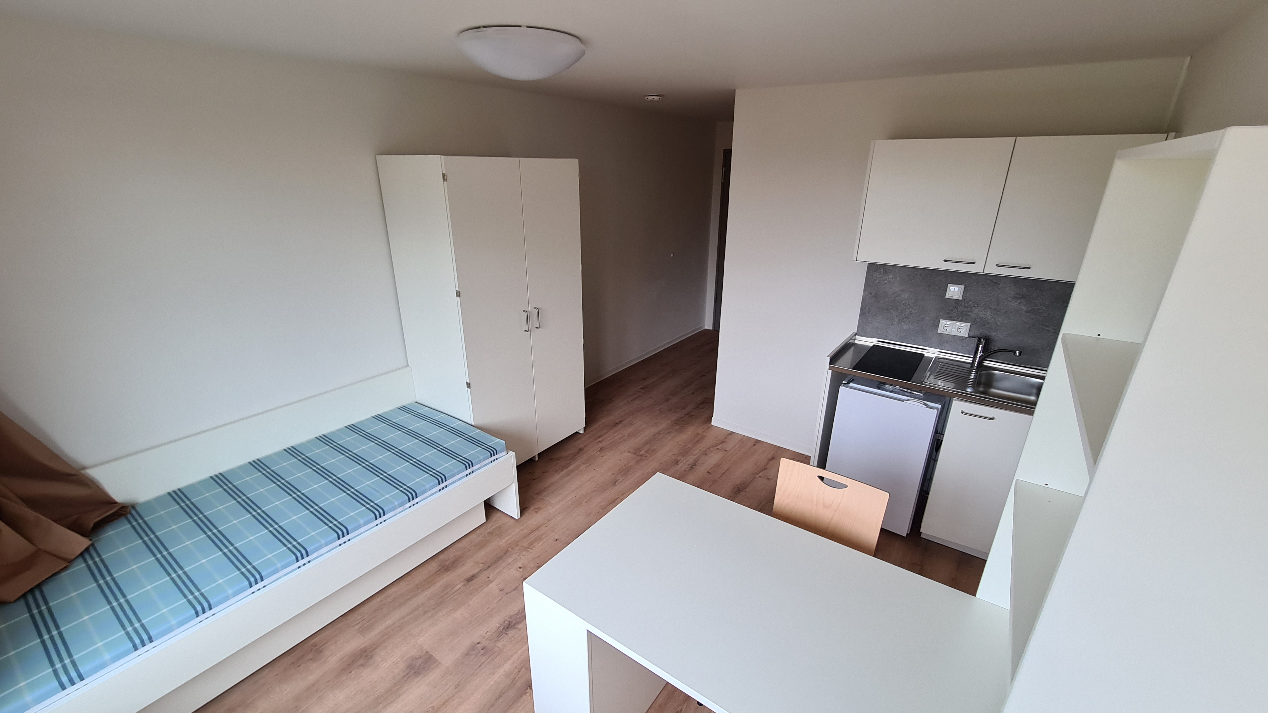 Zimmer mit Küchenzeile, Schrank, Regal, Tisch, Stuhl und Bett mit Blickrichtung zur Tür.