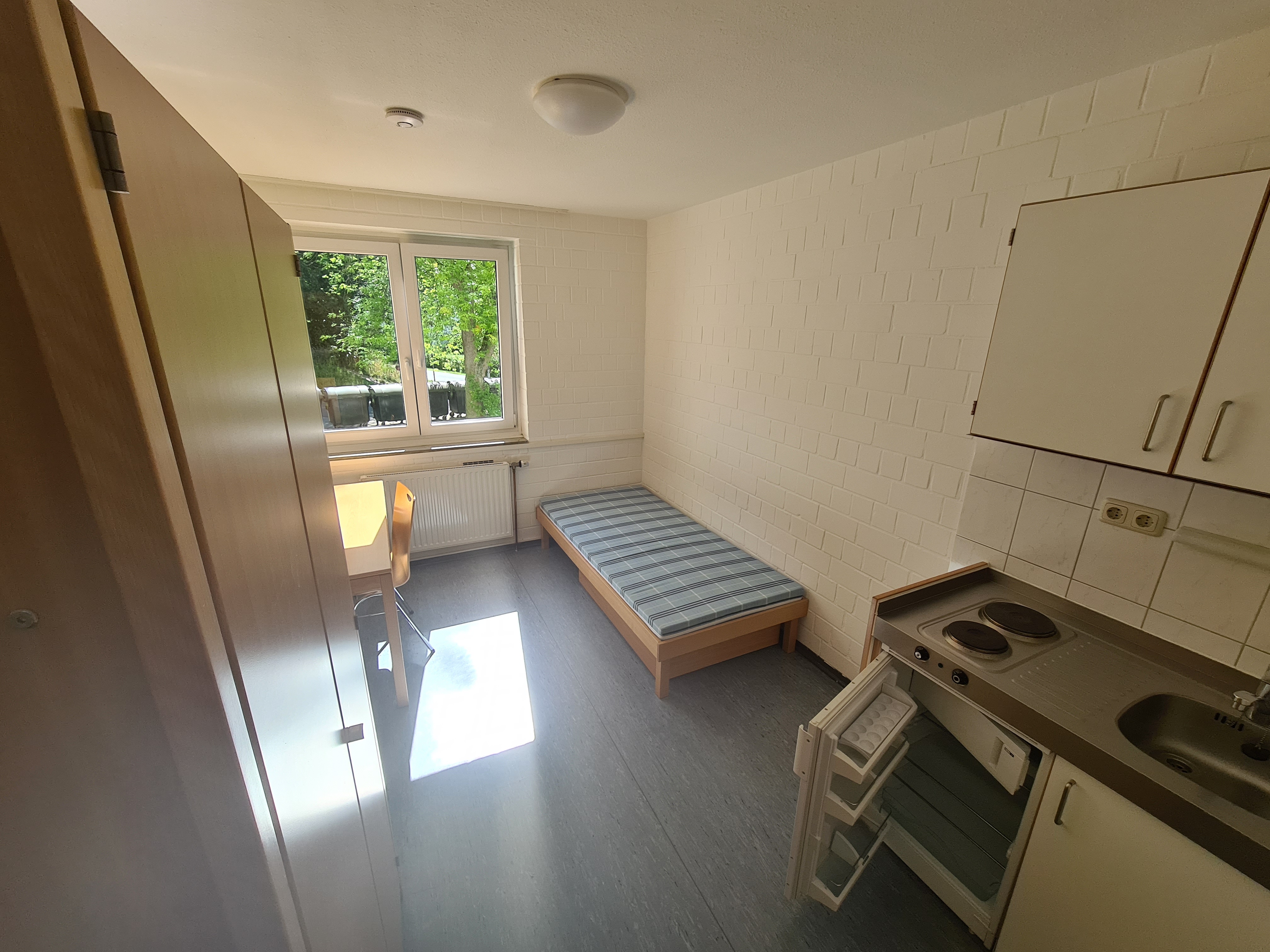 Zimmer mit Küchenzeile, Schrank, Tisch, Stuhl und Bett.